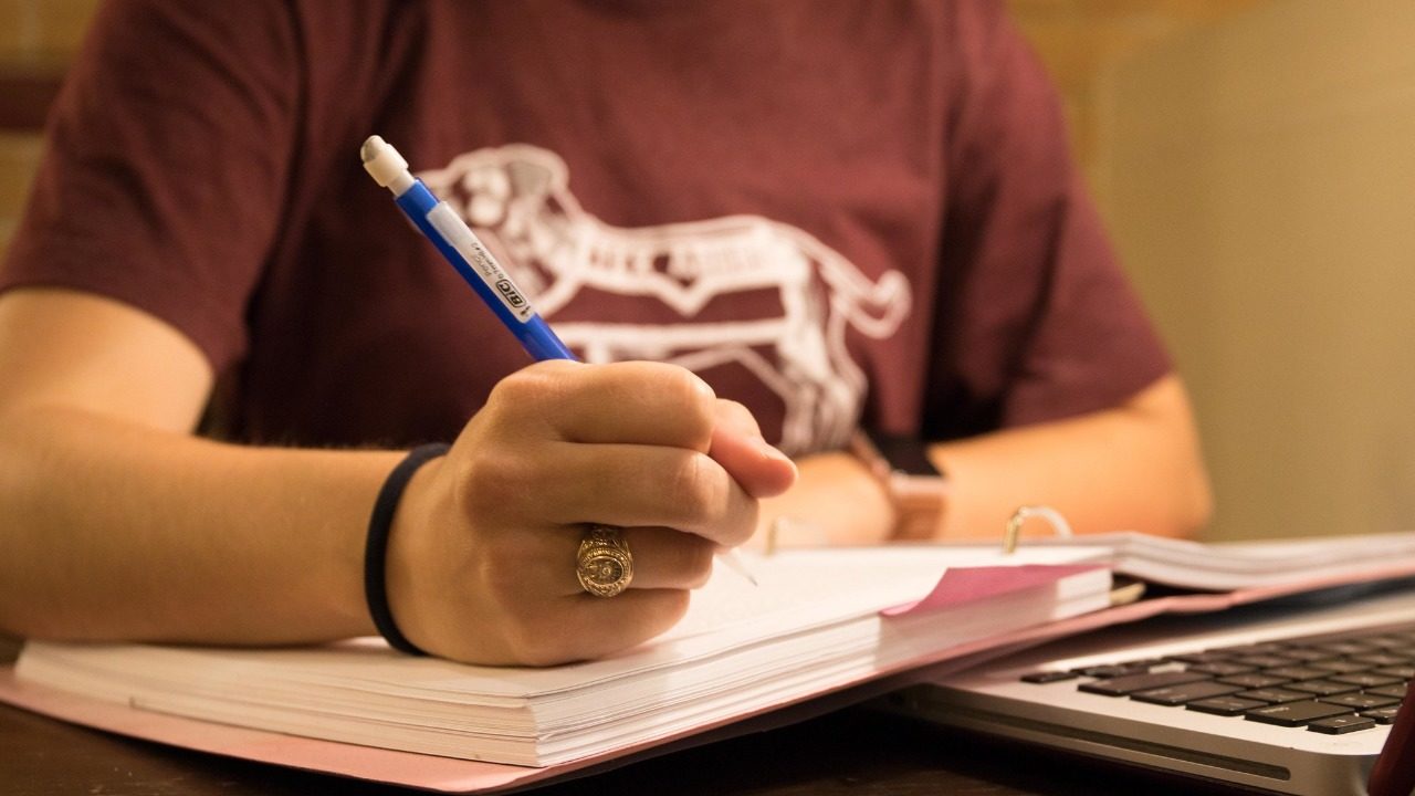Student, Ritika's hand in view holding a pencil writing on pages. In view you can see her Aggie Ring on her right ring finger
