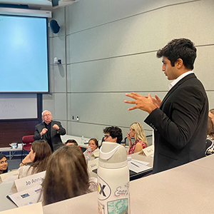 A student dressed in professional attire standing from the audience in class, talking with a professor at the front of the class.