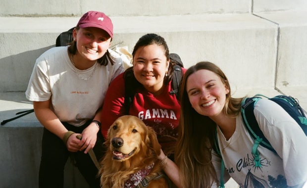 3 students sitting outdoors on a concrete step on a sunny day, smiling around a golden retriever dog sitting in the middle.