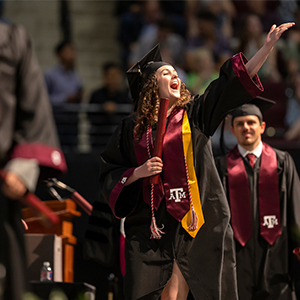 A student walking across the graduation stage smiling to the crowd and extending an arm in celebration.