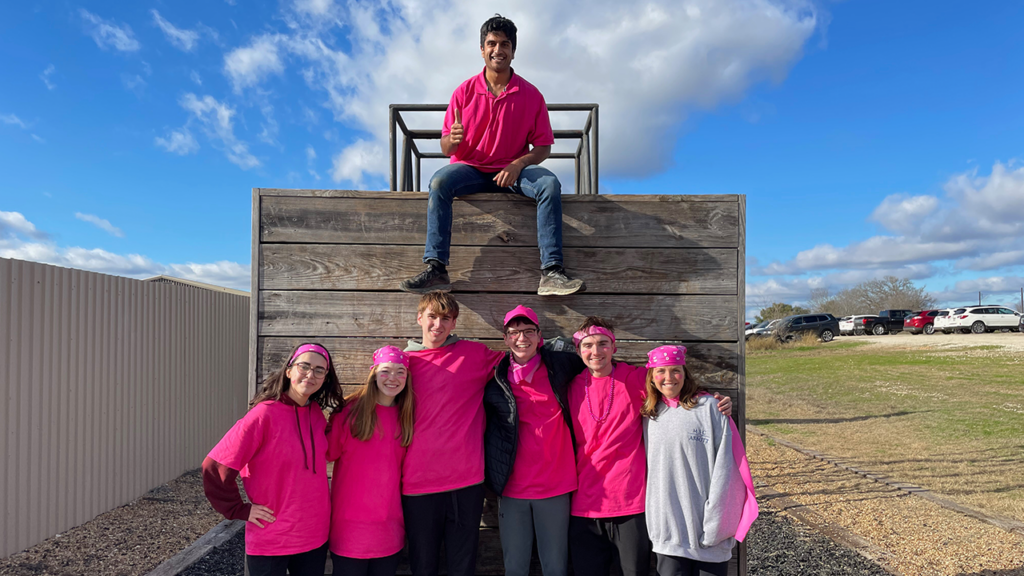 A group of friends dressed in pink shirts and bandanas, huddled together outside with a wooden wall in the background.