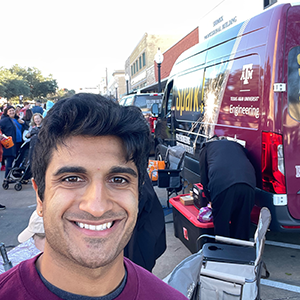 A student in the foreground of a selfie-style image outside with a Texas A&M Engineering Spark! van in the background