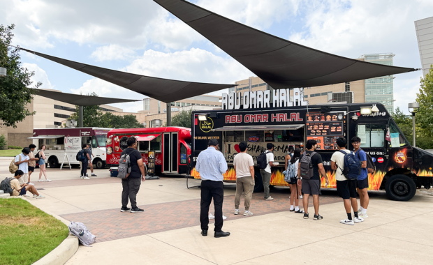 A food truck park outdoors on a cloudy day with three food trucks parked and people waiting in line.