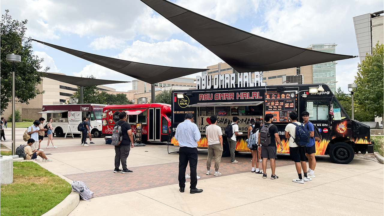 A food truck park outdoors on a cloudy day with three food trucks parked and people waiting in line.