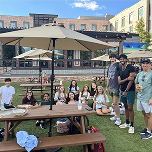 A group of students sitting behind a picnic table on green turf grass, smiling.