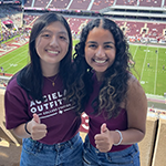 Two students standing in the stands with Kyle Field in the background, smiling, wearing Texas A&M attire, Gig'em gesture.