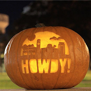 A pumpkin carved with the Texas A&M University skyline and "Howdy!" beneath, taken at night with a black sky in the background.