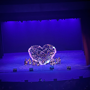 Taken from the view of the audience, a blue stage with many performers in the center creating a heart shape with their arms.